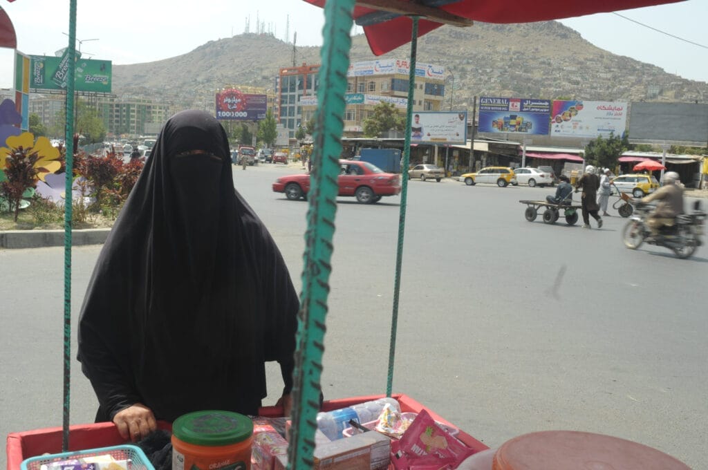 Women sometimes sold goods on the street - most professions are no longer accessible to women. Street vending is also becoming more and more restricted. A woman in a black niqab stands at a busy intersection in the city by a street cart filled with goods; hills and buildings can be seen in the background.