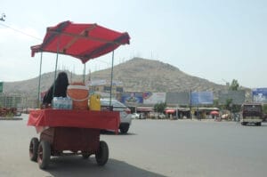 Ein Straßenverkaufswagen mit rotem Vordach und Wasserbehältern steht neben einer befahrenen Straße. Im Hintergrund ist ein Hügel mit Antennen und Gebäuden zu sehen.