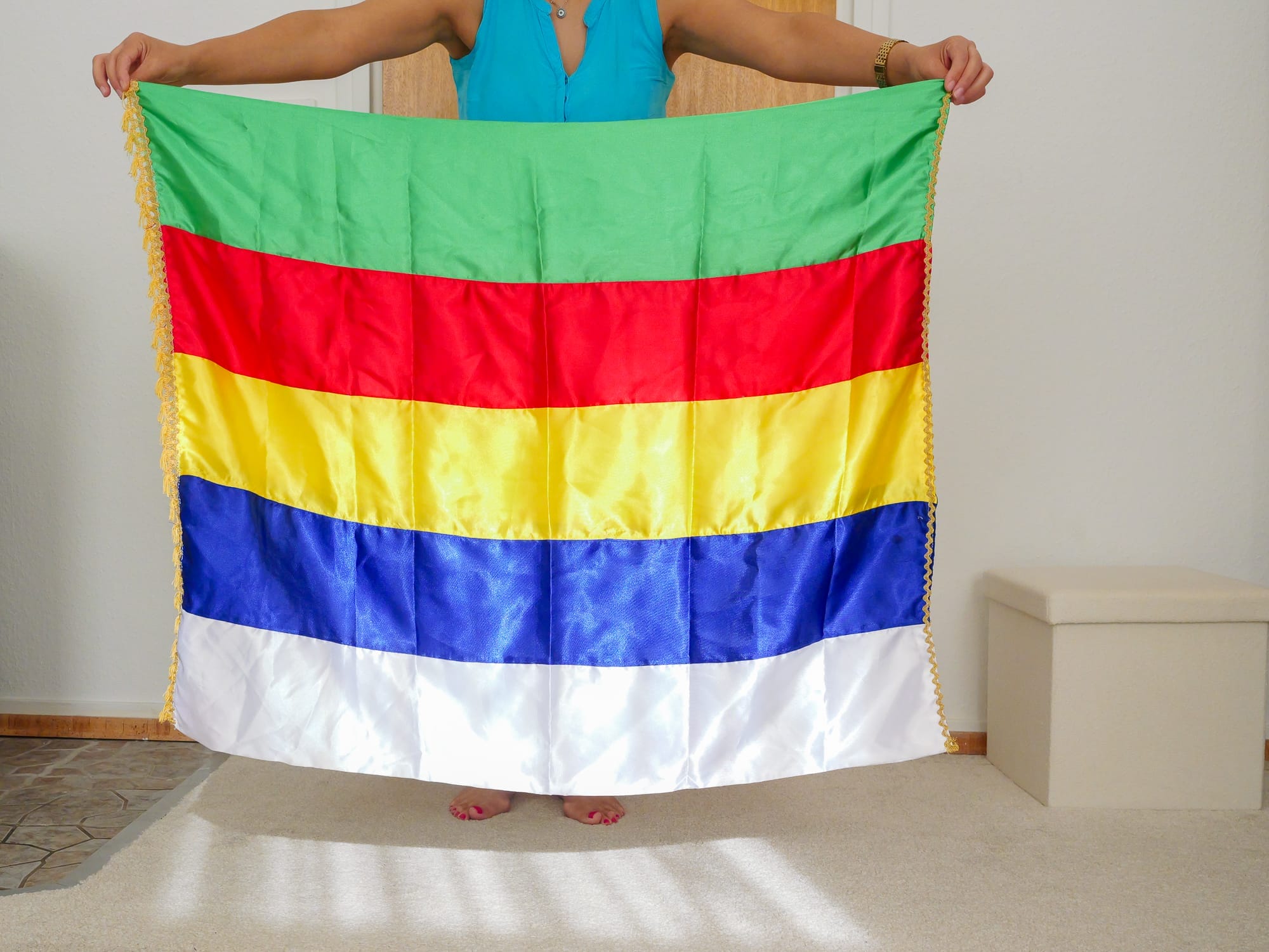 Eine Person hält eine Fahne mit horizontalen grünen, roten, gelben, blauen und weißen Streifen - Farben, die mit dem Erbe der Drusen assoziiert werden - und steht in einem Haus auf einem hellen Teppichboden. Diese Szene könnte aus as-Suweida stammen.