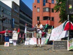 David Ginati spricht in einem Protestcamp im Freien mit Schildern, die auf einen Hungerstreik für Gaza hinweisen, in ein Mikrofon; im Hintergrund sind palästinensische Fahnen und Transparente zu sehen, die für Palästina eintreten.