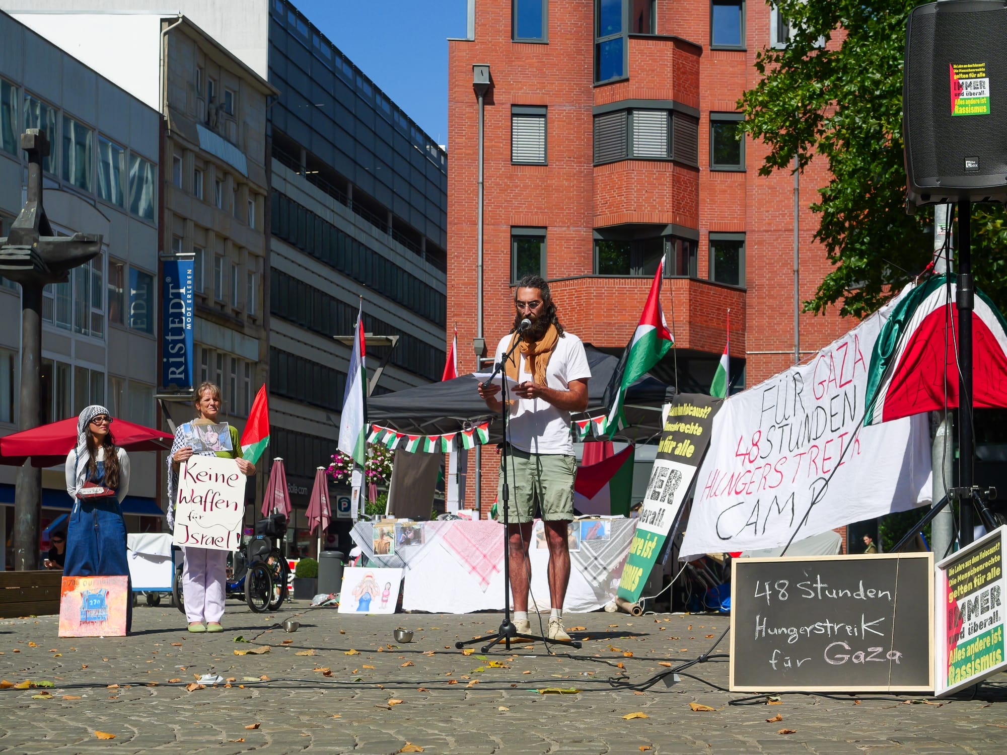 David Ginati spricht in einem Protestcamp im Freien mit Schildern, die auf einen Hungerstreik für Gaza hinweisen, in ein Mikrofon; im Hintergrund sind palästinensische Fahnen und Transparente zu sehen, die für Palästina eintreten.