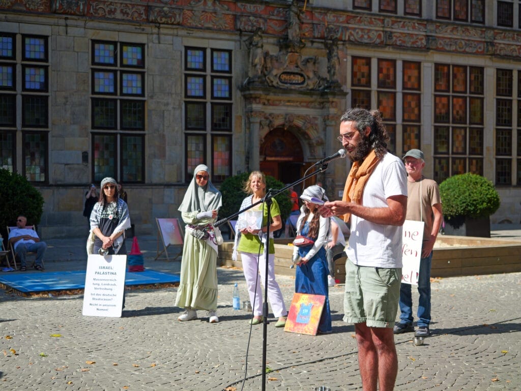 David Ginati spricht bei einer Versammlung im Freien für den Gazastreifen in ein Mikrofon. Hinter ihm stehen mehrere Personen mit Schildern und Plakaten vor einem historischen Gebäude.