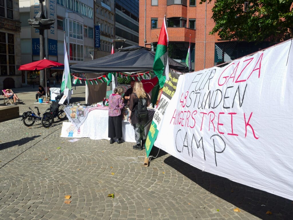 Protestcamp im Freien mit Zelten und Transparenten; auf einem großen Schild steht "Hilf Gaza, 48 Stunden Hungerstreik Camp". Menschen versammeln sich an den Tischen, Palästina- und Gaza-Fahnen sind zu sehen, als David Ginati sich zu den Unterstützern gesellt, um Solidarität zu zeigen.