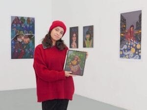 Eine Frau mit rotem Pullover und Mütze, möglicherweise Kimia, hält ein gerahmtes Kunstwerk in der Hand, während sie in einer Galerie steht, in der hinter ihr bunte Gemälde ausgestellt sind - eine Feier für kreative Werke in Deutschland.